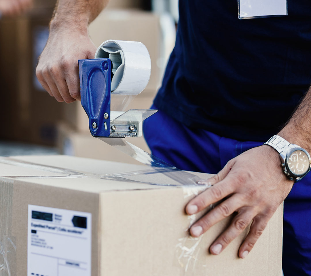 Close-up of delivery man closing carboard box with a tape while preparing packages for shipment.